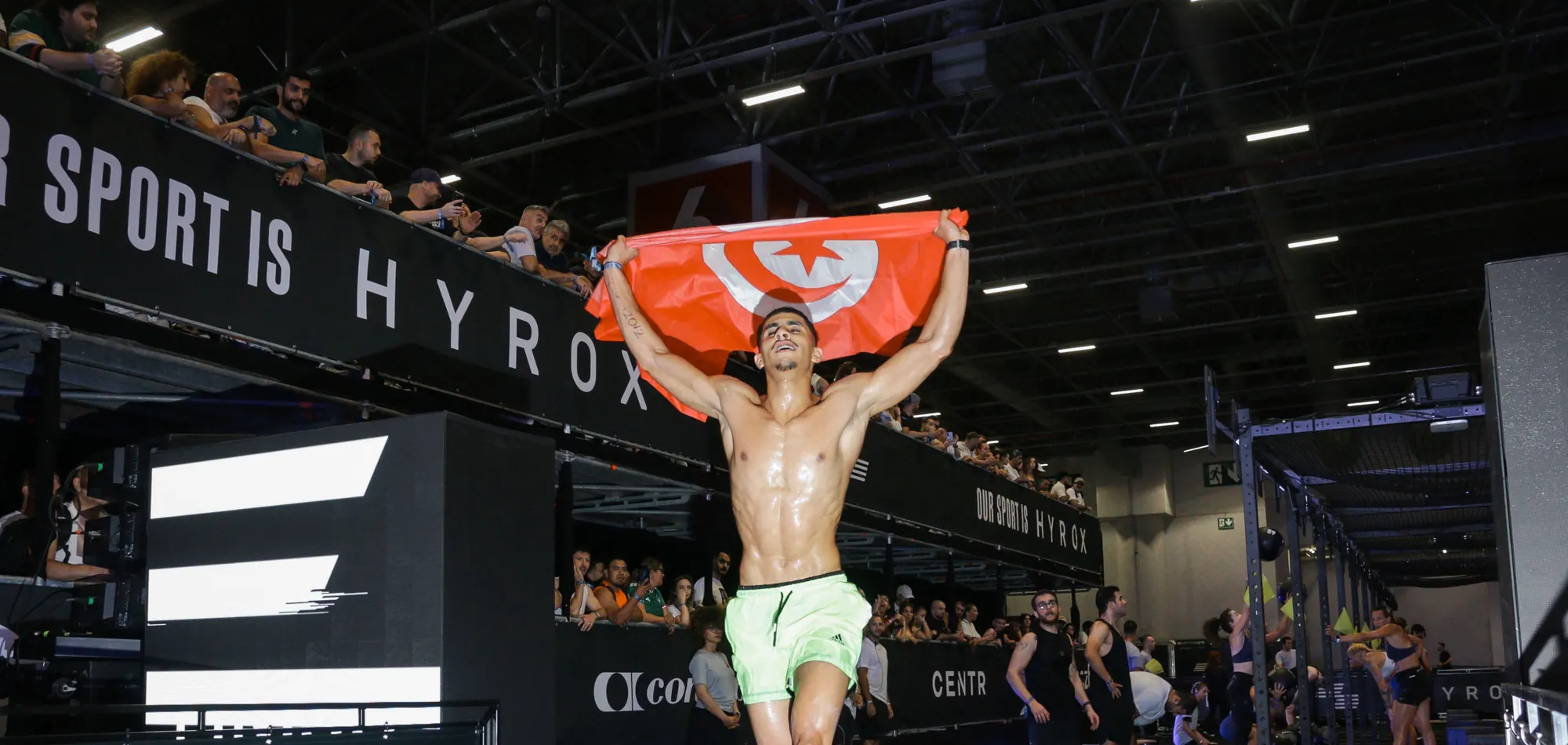 Athlete celebrating with a flag at the finish of a HYROX race in an exhibition hall